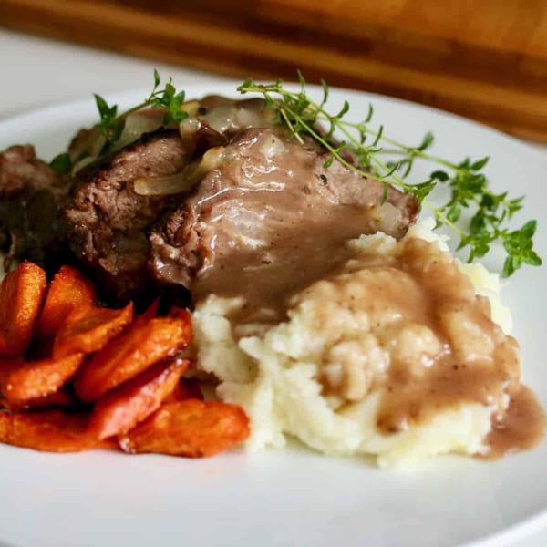 Braised beef with gravy, mashed potatoes, roasted carrots, and fresh thyme on a white plate.