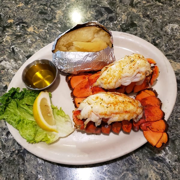 Two lobster tails with a foil-wrapped baked potato, butter, lettuce, and a lemon wedge.