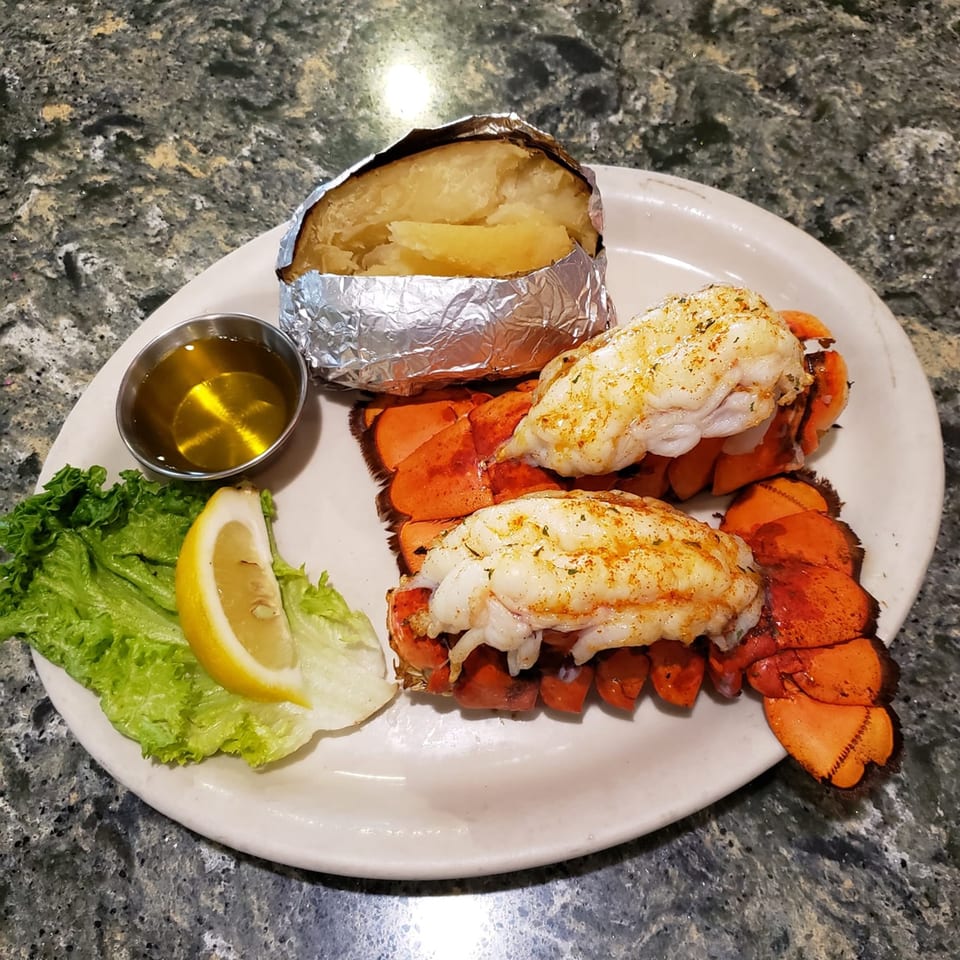 Two lobster tails with a foil-wrapped baked potato, butter, lettuce, and a lemon wedge.