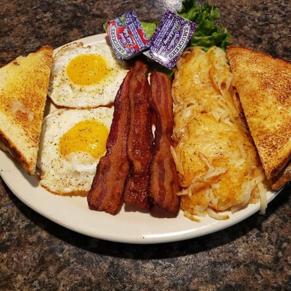 A breakfast plate with two fried eggs, crispy bacon, hash browns, toast, and jam packets.
