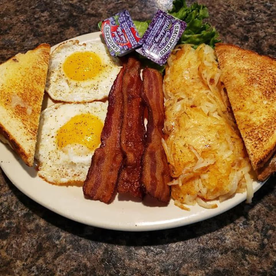 A breakfast plate with two fried eggs, crispy bacon, hash browns, toast, and jam packets.